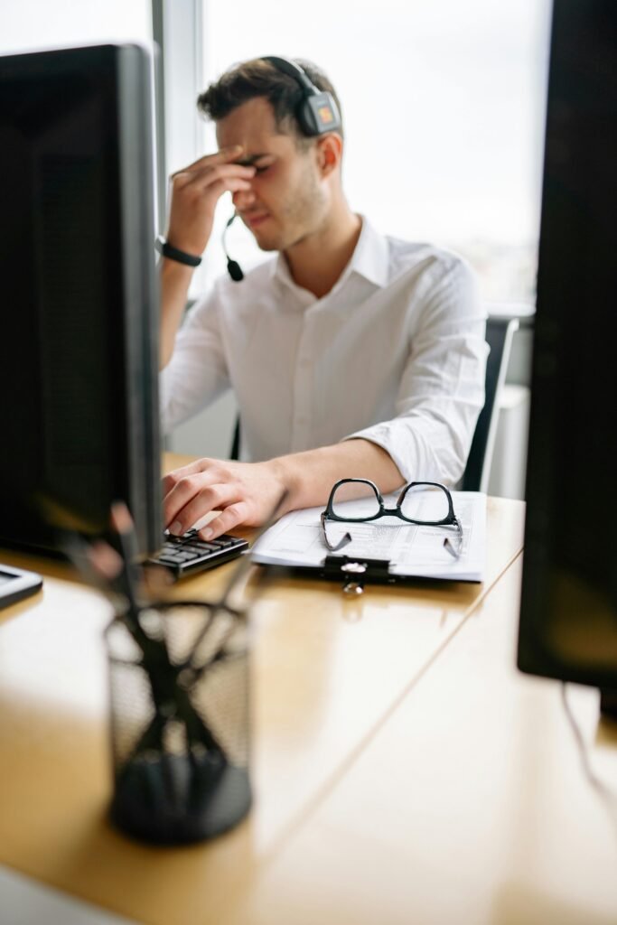 A tired call center employee with headset looks stressed at his office desk.